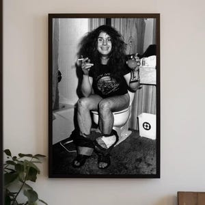 May include: Black and white photo of a person sitting on a toilet, smoking a cigarette and holding a glass of liquid. The person is wearing a black t-shirt with a graphic on it and jeans.