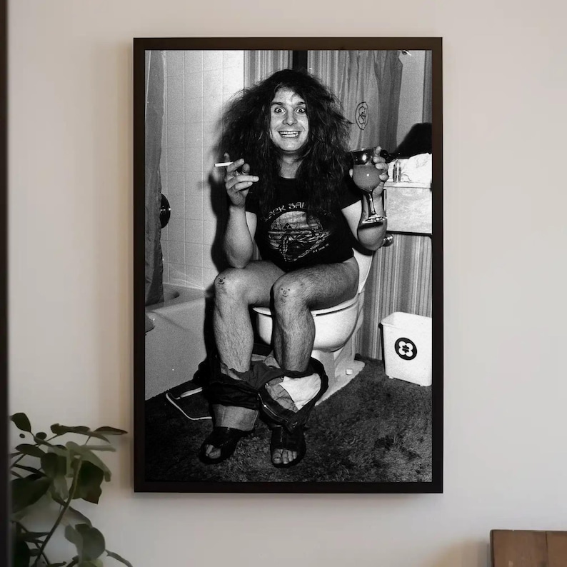 May include: Black and white photo of a person sitting on a toilet, smoking a cigarette and holding a glass of liquid. The person is wearing a black t-shirt with a graphic on it and jeans.