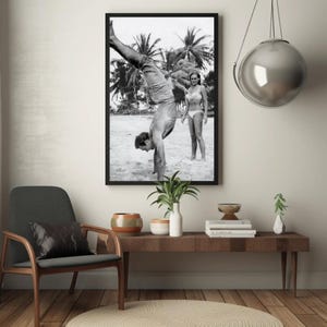 Op de afbeelding: Zwart-witfoto van een man die handstand maakt op een strand, met een vrouw die achter hem staat. De man draagt ​​een short en de vrouw een bikini. Palmbomen zijn zichtbaar op de achtergrond.