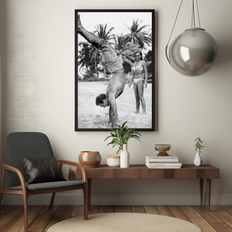 Op de afbeelding: Zwart-witfoto van een man die handstand maakt op een strand, met een vrouw die achter hem staat. De man draagt ​​een short en de vrouw een bikini. Palmbomen zijn zichtbaar op de achtergrond.