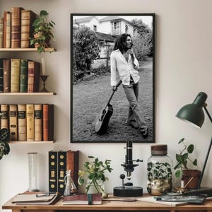 Op de afbeelding: Zwart-witfoto van een muzikant die een gitaar vasthoudt. De muzikant draagt een wit shirt en een spijkerbroek. De foto is buiten gemaakt en toont een huis op de achtergrond.