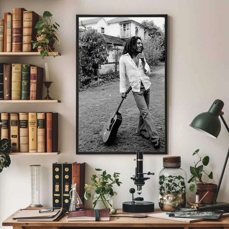 Op de afbeelding: Zwart-witfoto van een muzikant die een gitaar vasthoudt. De muzikant draagt een wit shirt en een spijkerbroek. De foto is buiten gemaakt en toont een huis op de achtergrond.