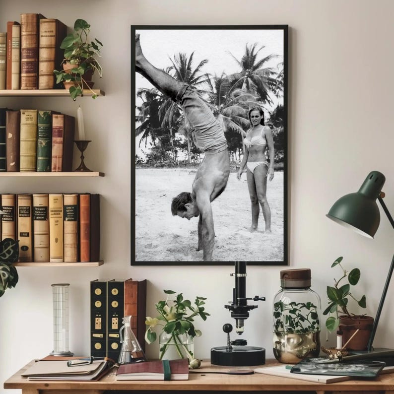 Op de afbeelding: Zwart-witfoto van een man die handstand maakt op een strand, met een vrouw die achter hem staat. De man draagt ​​zwembroek en de vrouw een bikini. De foto is ingelijst in een zwart frame.