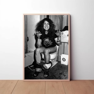 May include: Black and white photo of a man with long hair sitting on a toilet, smoking a cigarette and holding a glass of liquid. He is wearing a black t-shirt with a graphic on it. The photo is taken in a bathroom.