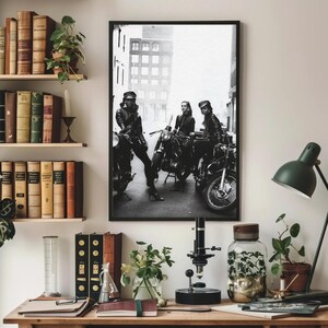 May include: Black and white framed print featuring three people posing with motorcycles on a city street. The image is displayed above a wooden desk with books, plants, and a microscope.