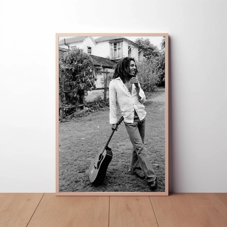 Op de afbeelding: Zwart-witfoto van een man met dreadlocks die een gitaar vasthoudt. Hij draagt een wit shirt en een spijkerbroek. De foto is gemaakt buiten voor een gebouw.