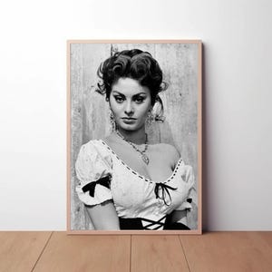 May include: Black and white portrait photograph of a woman with dark hair styled in a bouffant. She is wearing a white blouse with a low neckline and a black skirt. She is looking off to the side.