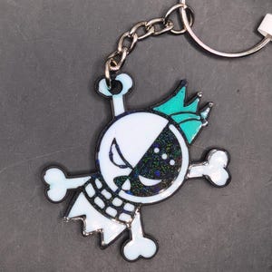 Puede incluir: Llavero novedoso con diseño de calavera dividida en dos, una mitad blanca y la otra negra con purpurina. La calavera tiene una corona turquesa y huesos cruzados. Incluye cadena y llavero plateado.