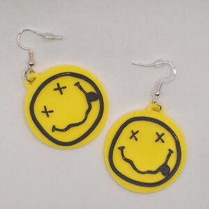 Puede incluir: Un par de pendientes amarillos con un diseño de cara sonriente negra. Los pendientes tienen ganchos plateados y forma circular. Cada cara sonriente tiene ojos tachados y una boca curvada.