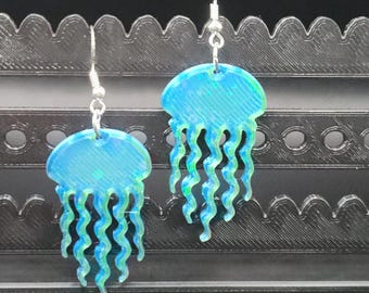 May include: A pair of jellyfish earrings. Each earring features a blue and green jellyfish design with wavy tentacles. The earrings are suspended from silver-colored hooks.