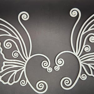 May include: A pair of white, decorative, plastic butterfly wings with intricate designs. The wings are connected by a heart shape.