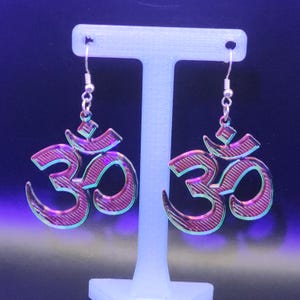 May include: A pair of Om symbol earrings. The earrings are a metallic pink with a striped pattern and a silver hook. The earrings are displayed on a white stand against a dark blue background.