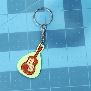 Puede incluir: Un llavero verde con forma de guitarra con un contorno marrón y la palabra "Bass" escrita en él. El llavero está unido a un llavero plateado.