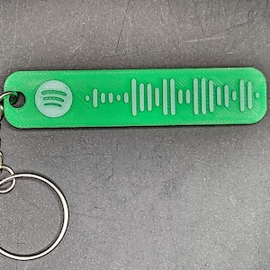 May include: A green rectangular keyring featuring a Spotify code design. The keyring has a silver chain and a round key ring. The Spotify logo is visible on the left. The background is a grey surface.