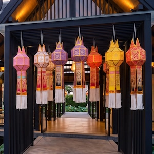 May include: A dark-roofed building with a covered walkway featuring rows of hanging cylindrical paper lanterns in shades of pink, orange, yellow, and purple, with white tassels, overlooking a pond filled with green lily pads and pink flowers.