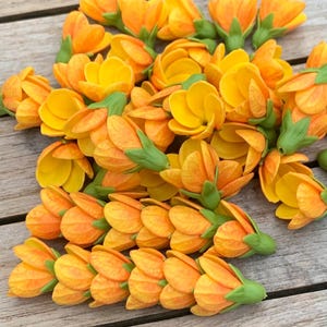 Puede incluir: Una colección de pequeñas flores artificiales hechas a mano en tonos amarillo brillante y naranja. Cada flor presenta pétalos en capas y un cáliz verde en la base. Algunas flores están dispuestas en grupos lineales, mientras otras están sueltas.