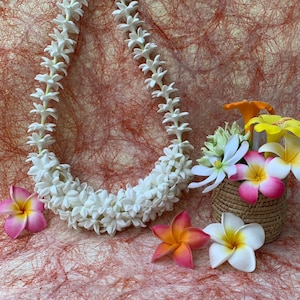 以下が含まれることがあります： 白い花のレイネックレスと、カラフルなプルメリアの花。 花はピンク、オレンジ、黄色、白で、質感のある赤茶色の表面に配置されています。 小さな織りのバスケットには、追加の花が入っています。