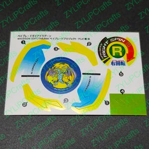 Könnte beinhalten: Ein weißer Aufkleberbogen mit Anweisungen zum Zusammenbauen eines Beyblade-Spielzeugs. Der Bogen ist in den Farben Gelb, Blau und Grün gehalten. Der Aufkleberbogen enthält ein Bild des Beyblades, einem Kreiselspielzeug. Der Text auf dem Aufkleberbogen lautet "Beyblade Gaia Dragoon" und "Right Spin".