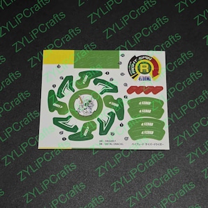 Può includere: Un foglio di adesivi per un giocattolo Beyblade, con disegni verdi e bianchi. Gli adesivi includono varie forme, testo e un emblema circolare con le parole "RIGHT SPIN" e "右回転". Il testo "AR-DRIVER F" e "METAL DRACIEL" è anche visibile.