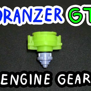 May include: A lime green and gray toy component with the text "DRANZER GT" and "ENGINE GEAR". The toy has a multi-piece design with a central gray section and a lime green top. The background is black.