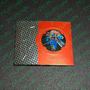 May include: A red and black card with a circular image of a character from a video game. The character is wearing a blue and orange outfit and holding a sword. The text "サイゾー" is printed on the card.