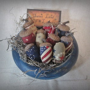 May include: A blue bowl filled with fabric-covered acorns decorated with red, white, and blue patriotic colors. The bowl is surrounded by dried twigs and a sign that reads "Sweet Land of Liberty".