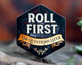 May include: A black and white hexagonal sticker with the text "ROLL FIRST ASK QUESTIONS LATER" in white and gold. The sticker is set against a blurred background with a rock and foliage.