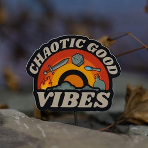 May include: A sticker with the words "CHAOTIC GOOD VIBES" in a stylized design. The sticker features a rainbow, a sword, a die, a potion bottle, and flames. The background is a blurred blue and brown.