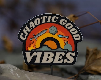 May include: A sticker with the words "CHAOTIC GOOD VIBES" in a stylized design. The sticker features a rainbow, a sword, a die, a potion bottle, and flames. The background is a blurred blue and brown.