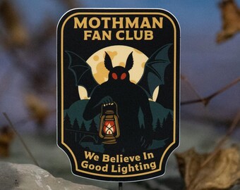 May include: A black and gold Mothman Fan Club sticker featuring a silhouette of the Mothman holding a lantern, with the text "We Believe In Good Lighting". The design includes a full moon and bat wings.