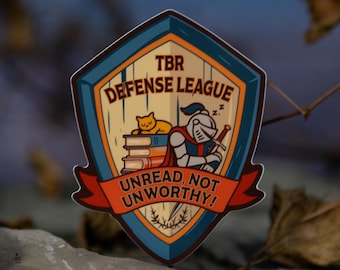 May include: A shield-shaped sticker with a cartoon design. The sticker features a sleeping knight, a cat, and the text "TBR Defense League: Unread Not Unworthy!" in a red banner. The sticker has a blue and orange color scheme.