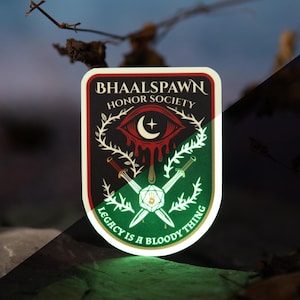 May include: A sticker with the text "BHAALSPAWN HONOR SOCIETY" and the phrase "LEGACY IS A BLOODY THING." The sticker features a red eye with a crescent moon, crossed swords, and a green and white design. Two dice are in the background.