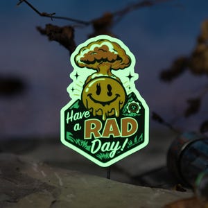 Może przedstawiać: Świecąca naklejka z uśmiechniętą twarzą i wzorem grzyba atomowego. Naklejka głosi "Have a RAD Day!" w kolorze zielonym i białym. Naklejka znajduje się na ciemnym tle z liśćmi i skałą.
