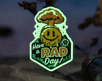 May include: A glowing sticker with a smiling face and a mushroom cloud design. The sticker reads "Have a RAD Day!" in green and white. The sticker is on a dark background with leaves and a rock.