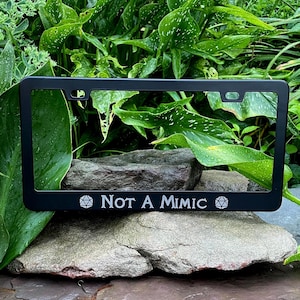 May include: Black license plate frame with the text "Not A Mimic" in white. The frame has a rectangular shape with rounded corners and is set against a backdrop of green foliage and rocks. The text is in a bold, sans-serif font.