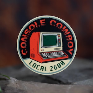 May include: A round sticker featuring a retro computer design in red, cream, and black. The text "CONSOLE COWBOY" arcs around the top, with "LOCAL 2600" at the bottom. The sticker has a vintage, tech-inspired aesthetic.