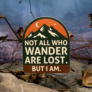 Può includere: Un adesivo rettangolare con un disegno di catena montuosa in verde, arancione e bianco, con una luna crescente e una stella. Il testo recita: «NOT ALL WHO WANDER ARE LOST. BUT I AM.»