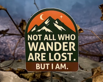 May include: A rectangular sticker with a mountain range design in green, orange, and white, with a crescent moon and star. The text reads, "NOT ALL WHO WANDER ARE LOST. BUT I AM."