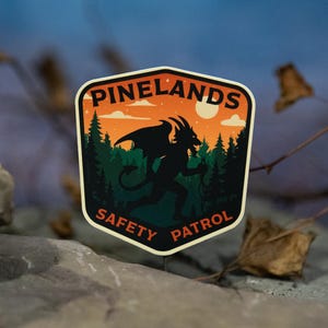 May include: A sticker with a shield shape featuring the text "PINELANDS SAFETY PATROL". The design includes a silhouette of a horned creature with wings, trees, and a sunset. The sticker is set against a blurred background.
