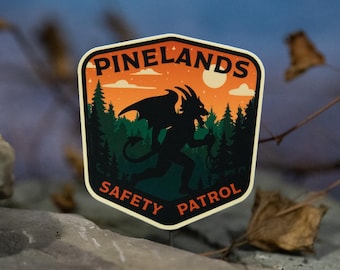 May include: A sticker with a shield shape featuring the text "PINELANDS SAFETY PATROL". The design includes a silhouette of a horned creature with wings, trees, and a sunset. The sticker is set against a blurred background.