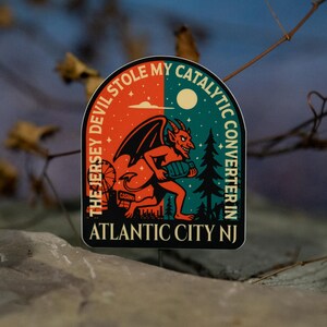 May include: A sticker with a devilish cartoon character holding a catalytic converter. The text reads "The Jersey Devil Stole My Catalytic Converter In Atlantic City NJ." The background is split between orange and teal.