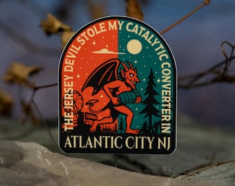 May include: A sticker with a devilish cartoon character holding a catalytic converter. The text reads "The Jersey Devil Stole My Catalytic Converter In Atlantic City NJ." The background is split between orange and teal.