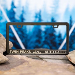 May include: A black license plate frame with the text "TWIN PEAKS AUTO SALES" in white. The frame has a rectangular shape with rounded corners and is displayed against a blurred blue and white background.