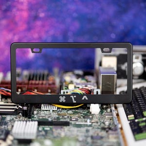 May include: Black license plate frame with white text and a logo. The frame is positioned in front of a computer motherboard and keyboard, with a colorful galaxy-themed background.