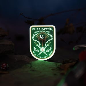 Puede incluir: Un parche verde y blanco con el texto "BHAALSPAWN HONOR SOCIETY" y "LEGACY IS A BLOODY THING". El parche presenta un ojo, una luna creciente, espadas y un dise&ntilde;o de corona. El parche est&aacute; iluminado con un brillo verde.