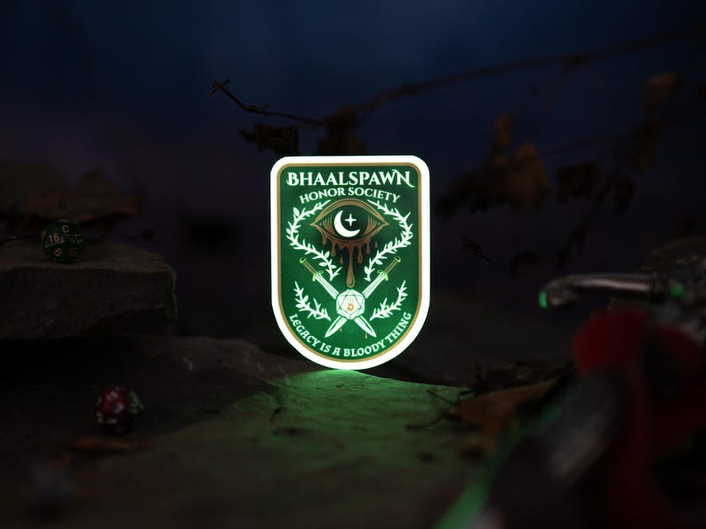Puede incluir: Un parche verde y blanco con el texto "BHAALSPAWN HONOR SOCIETY" y "LEGACY IS A BLOODY THING". El parche presenta un ojo, una luna creciente, espadas y un dise&ntilde;o de corona. El parche est&aacute; iluminado con un brillo verde.
