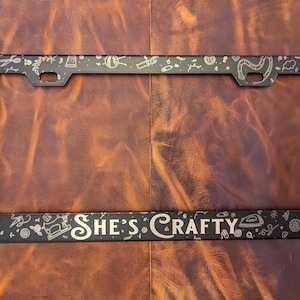 May include: Black metal license plate frame with a white outline and the text "She's Crafty" in a decorative font. The frame is decorated with small, white, craft-themed illustrations.