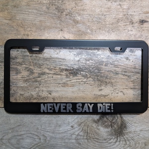 Puede incluir: Marco de matrícula negro con el texto "NEVER SAY DIE!" en letras grises. El marco tiene forma rectangular con esquinas redondeadas y recortes para el montaje. El marco es de metal.