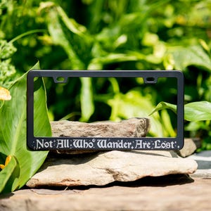 Not all who wander are lost fantasy literary quote engraved on a black stainless steel license plate frame on a natural mountain and vegetation themed background