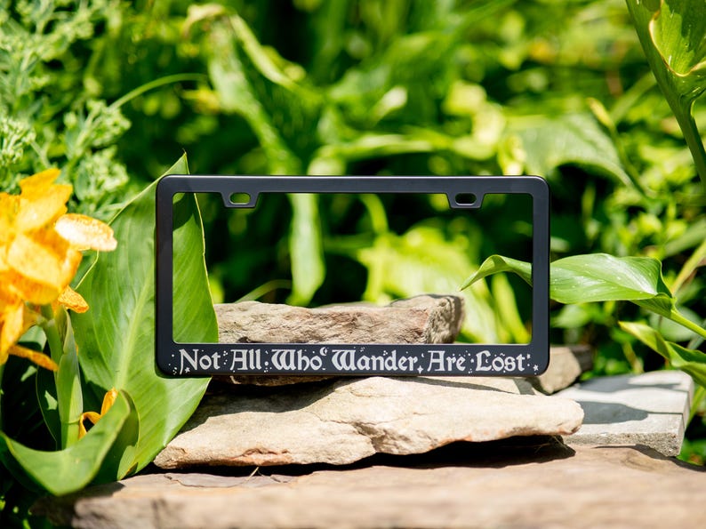 Not all who wander are lost fantasy literary quote engraved on a black stainless steel license plate frame on a natural mountain and vegetation themed background